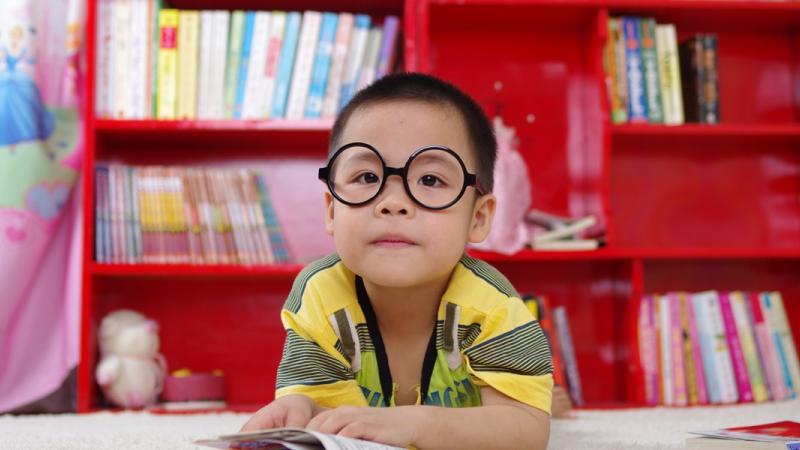 Child with glasses facing camera posed in front of a red bookshelf full of children's books.